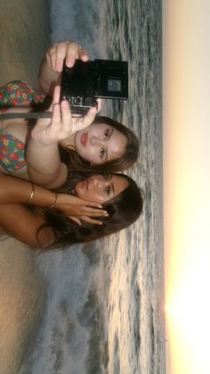 two young women standing next to each other on the beach taking pictures with their cell phones