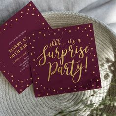 two red and gold surprise party cards sitting on top of a white plate with greenery