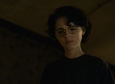 a woman wearing glasses standing in the dark with her head turned to the side and looking off into the distance