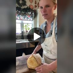 a woman in an apron holding a loaf of bread on top of a wooden table