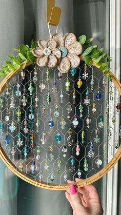 a person holding up a wooden frame with beads and leaves on it in front of a window