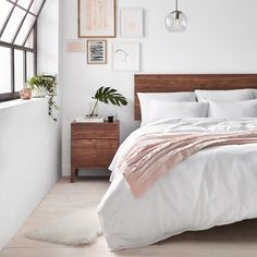 a bed with white sheets and pillows in a bedroom next to a window, potted plant and framed pictures on the wall