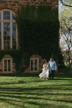 a bride and groom walking through the grass in front of an old brick building with ivy growing on it