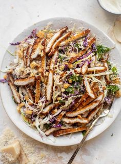 a white plate topped with chicken and coleslaw next to a bowl of dressing