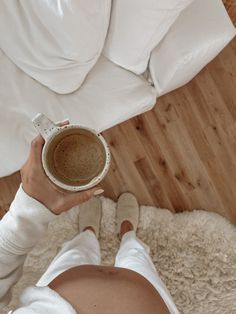 a woman holding a cup of coffee on top of a white rug next to a bed