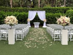 an outdoor wedding setup with white chairs and flowers on the aisle, surrounded by greenery