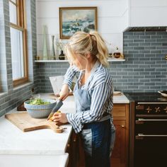 a woman standing in a kitchen preparing food on top of a wooden cutting board next to a window