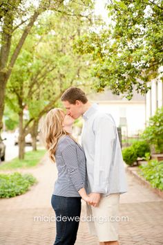 an engaged couple kissing in the middle of a brick walkway surrounded by trees and greenery