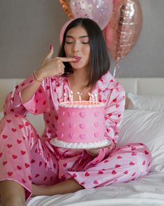 a woman in pink pajamas sitting on a bed with a cake and balloons behind her