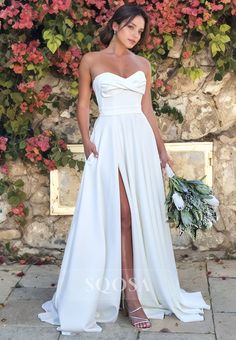 a woman wearing a white dress standing in front of a wall with flowers and greenery