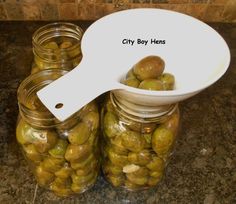 some jars filled with green olives sitting on top of a counter next to a white spoon