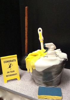 there is a cake that looks like a bucket with cleaning supplies on it and a sign next to it