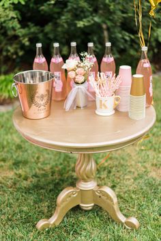 a table topped with lots of pink and gold cups filled with drinks on top of green grass