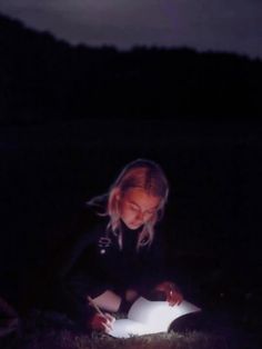 a woman sitting in the grass reading a book at night with her flashlight lit up