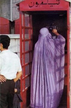 a woman in a purple shawl taking pictures from a red phone booth while another man looks on