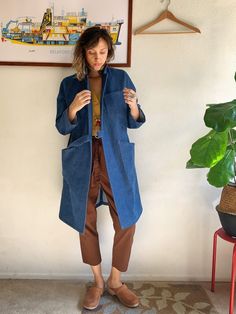 a woman standing in front of a wall wearing a blue coat and brown pants with her hands on her hips