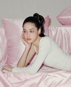 a woman laying on top of a bed next to pink sheets and pillows with her hand under her chin