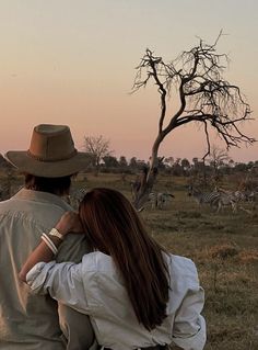 a man and woman standing in front of a tree with zebras behind them at sunset