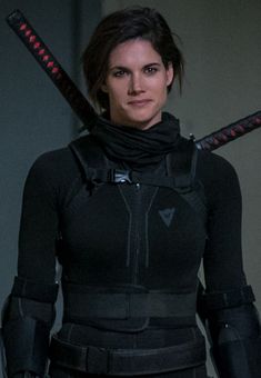 a woman with two swords in her hand and wearing a black outfit, standing next to a wall