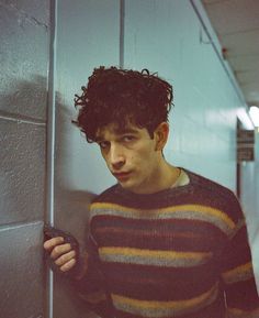 a young man leaning against a wall holding a cell phone in his right hand and looking at the camera