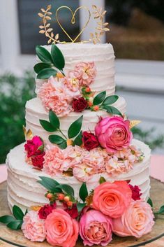 a wedding cake with pink flowers and greenery on the top is topped with a gold heart