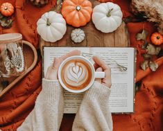 a person holding a cup of coffee in front of an open book and pumpkins