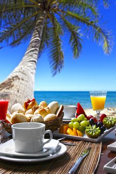 a table with plates and cups filled with food on it next to the ocean, under a palm tree