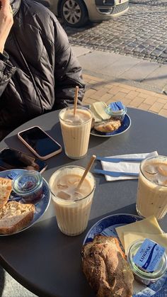 a man sitting at an outdoor table with two cups of coffee and sandwiches on it