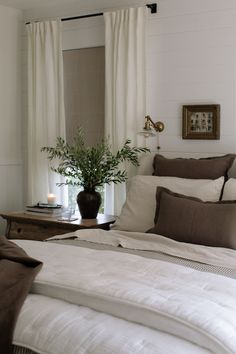 a bed with white sheets, pillows and a plant in a vase on the end table