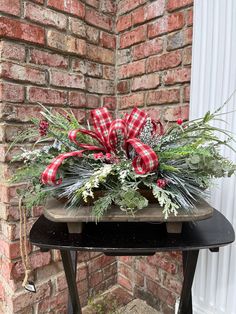 a wooden table topped with greenery and plaid bow on it's top, next to a brick wall