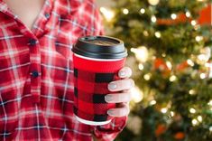 a woman holding a red plaid coffee cup in front of a christmas tree