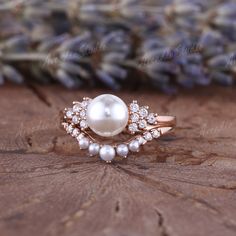 a pearl and diamond ring sitting on top of a piece of wood with lavender flowers in the background