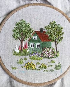 a green house surrounded by flowers and trees on a white linen covered surface with a wooden hoop