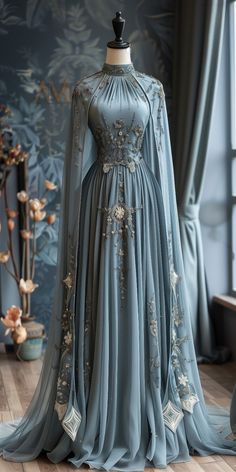 **A light blue evening gown with a cape. Embroidered details on the gown. A product photo showing the gown on a mannequin in a luxury boutique. Medieval Evening Gown, Fae Ballgown, Clothes On Mannequin, Blue Queen Dress, Light Blue Evening Gown, Wide Sleeve Dress, Otto Hightower, Dress Medieval, Blue Evening Gowns