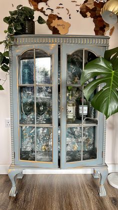 an old china cabinet with glass doors and gold trimmings on the top, in front of a large potted plant