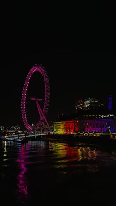 a ferris wheel lit up in the night sky over a body of water with buildings behind it