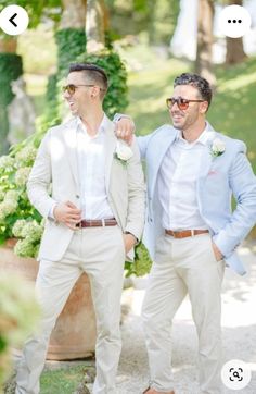 two men standing next to each other in front of trees and bushes, one wearing a light blue suit