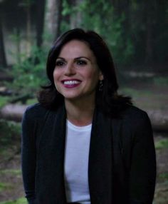 a woman standing in the woods with trees behind her and smiling at the camera while wearing a black blazer