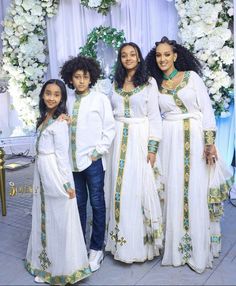 three women in white dresses standing next to each other near a flower covered wall with flowers on it