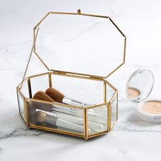 an open glass box with makeup brushes inside on a marble countertop next to a compact mirror