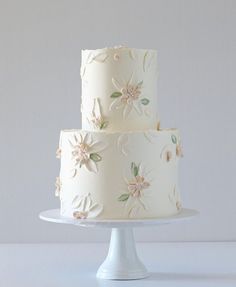 a three tiered cake with white frosting and flowers on the top is sitting on a pedestal