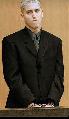 a man in a black suit and tie standing next to a wooden paneled wall