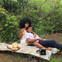 a man and woman sitting on a bench in the woods with food, drinks and snacks