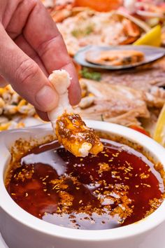 a person dipping some food into a white bowl with sauce on the table next to other foods