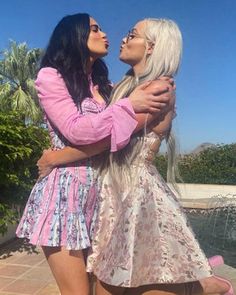 two women are hugging each other in front of a fountain with palm trees behind them