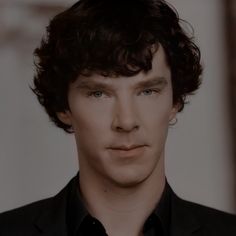 a young man with curly hair wearing a suit and black shirt looking at the camera