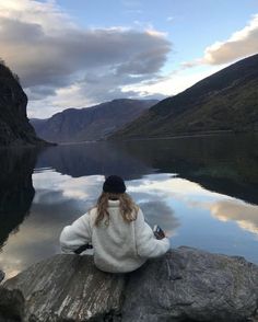 a woman sitting on top of a rock next to a body of water with mountains in the background