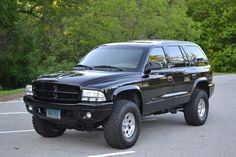 a large black truck parked in a parking lot next to some trees and bushes on the other side of the road