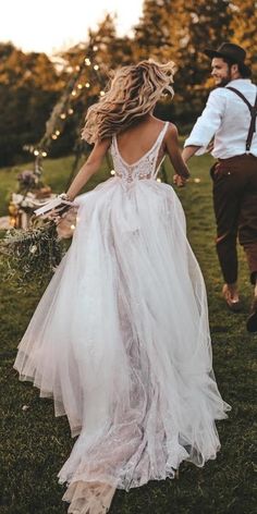 a man and woman walking in the grass holding hands, dressed in white dress with open back