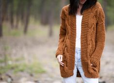 a woman standing in the woods wearing a brown knitted cardigan sweater and white jeans